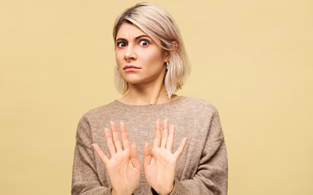 Женщина с короткими светлыми волосами поднимает ладони в жесте отказа на однотонном фоне